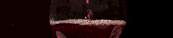 A stemmed glass being filled as red wine pours, sparkling in dark, moody lighting.
