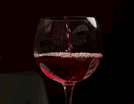 A stemmed glass being filled as red wine pours, sparkling in dark, moody lighting.