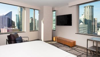 A modern hotel room with a bed, a chair, a wall-mounted TV, and floor-to-ceiling windows showcasing a city skyline with tall buildings.