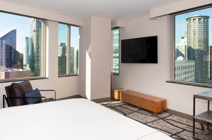 A modern hotel room with a bed, a chair, a wall-mounted TV, and floor-to-ceiling windows showcasing a city skyline with tall buildings.