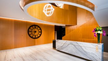 A modern lobby with a large clock on the wooden wall, a curved staircase, and a marble reception desk decorated with flowers.