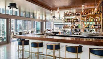 A modern bar with a long counter, stylish bar stools, and a fully stocked shelf of liquor bottles, located in a high-rise building with large windows.