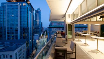 A modern urban balcony at dusk overlooks a city street lined with tall buildings. The balcony has seating and tables, with glass doors open inside.