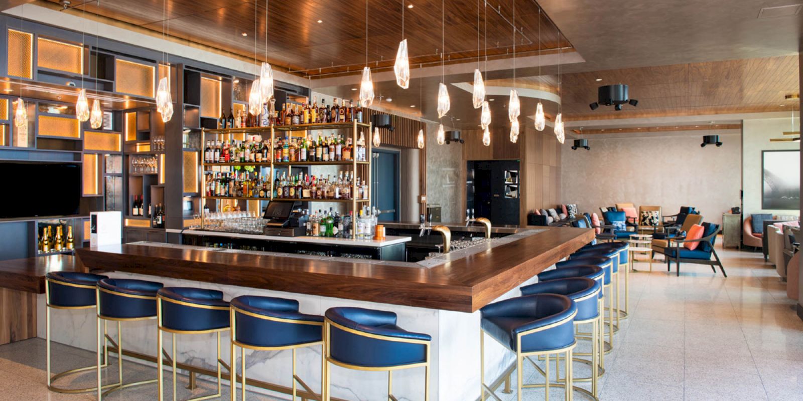 The image shows a modern bar with a wooden counter, blue bar stools, overhead lighting, and a fully stocked back shelf in a spacious, stylish setting.