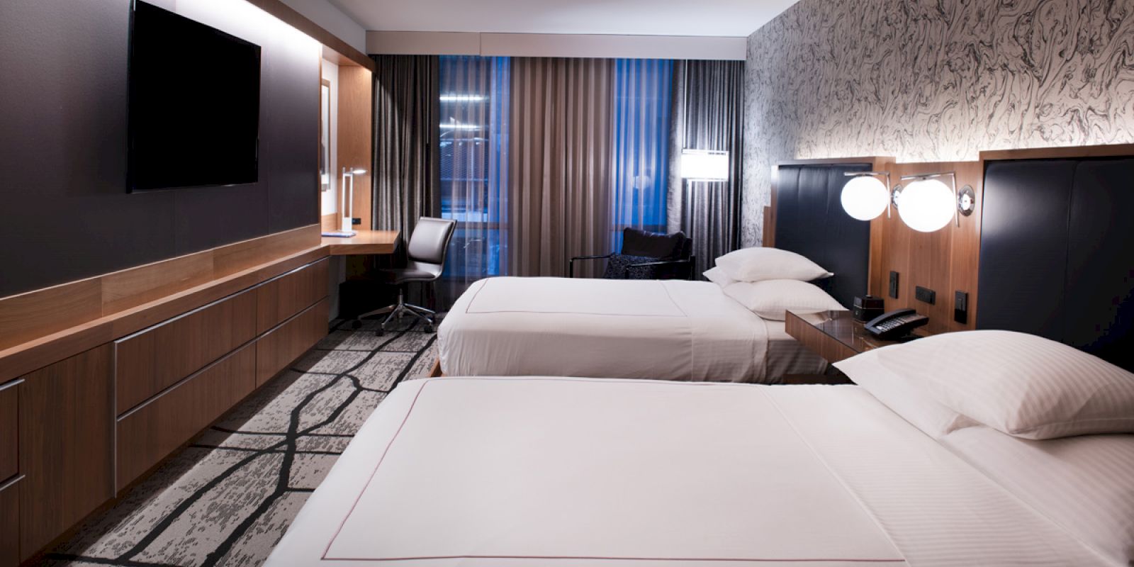 The image shows a modern hotel room with two neatly made beds, a wall-mounted TV, a desk with a chair, bedside tables, and contemporary decor.