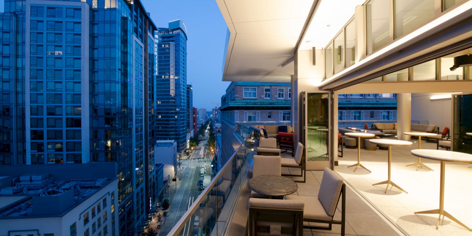 A modern balcony at dusk with a city view featuring tall buildings; the balcony has seating arrangements and tables near glass doors ending the sentence.