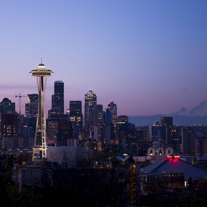 The image shows a twilight cityscape with a prominent tower (the Space Needle) and mountains in the background, likely depicting Seattle, Washington.