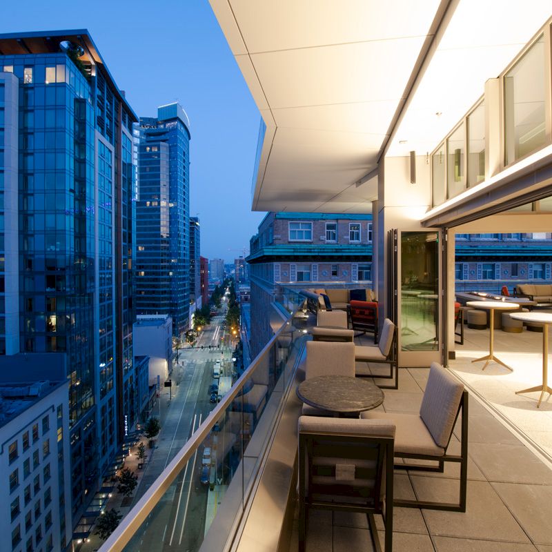 An urban evening scene with tall buildings and a modern balcony featuring outdoor furniture and tables, overlooking a city street.