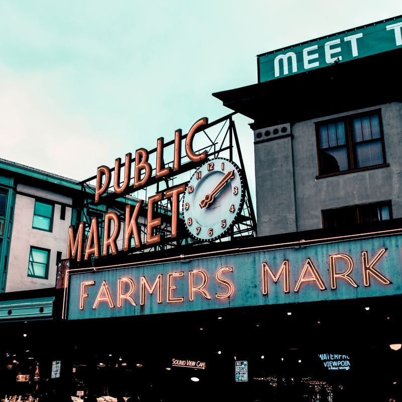 The image shows a neon-lit sign for a public farmers market, likely Pike Place Market in Seattle, with surrounding buildings and a clock.