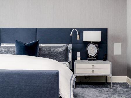 The image shows a contemporary bedroom with a blue and white color scheme, featuring a bed, nightstand with a lamp, decor item, and a sconce light fixture.