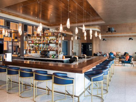 A modern bar with a well-stocked liquor display, blue chairs, and stylish pendant lights, surrounded by a cozy lounge area in a spacious room.