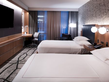 The image shows a modern hotel room with two beds, a flat-screen TV, a desk with a chair, and stylish decor illuminated by various light sources.