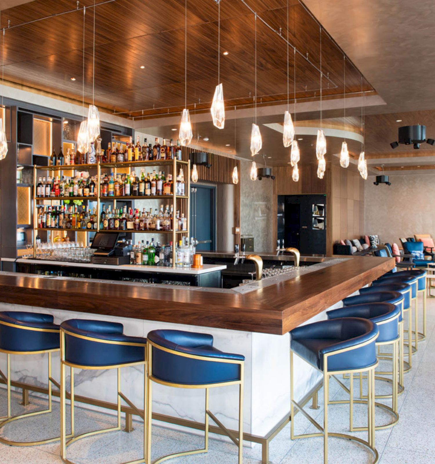 The image shows a modern bar with a central counter, blue and gold stools, hanging lights, and shelves stocked with various bottles.