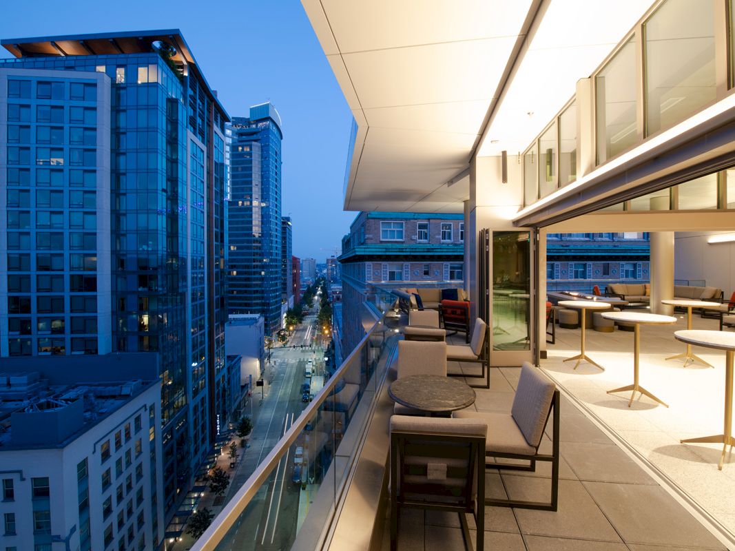 The image shows a modern rooftop terrace overlooking a cityscape with tall buildings. The area is furnished with tables and chairs.
