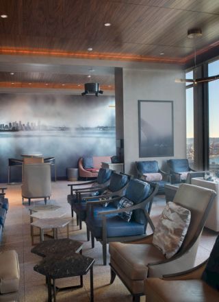 A modern lounge with plush seating, a cityscape wall mural, large windows showcasing a waterfront view, and chairs arranged around small tables.