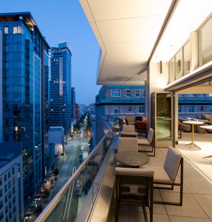 A modern cityscape view from a high-rise balcony at dusk, featuring outdoor seating and tables overlooking illuminated urban buildings and streets.