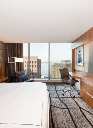 A modern hotel room with a large bed, desk, chair, and a big TV. The room has a large window with a city and water view.
