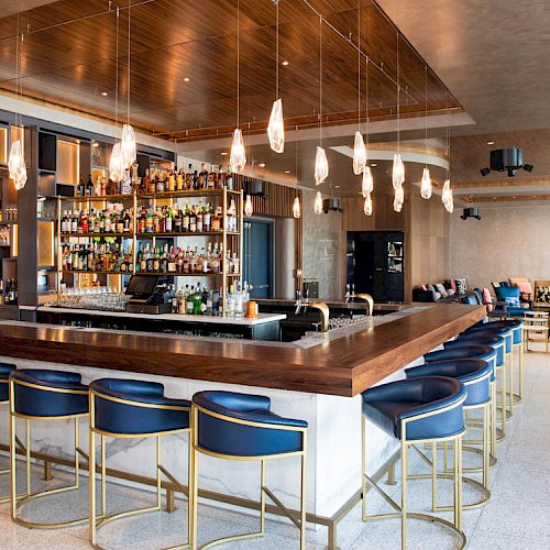 The image shows a modern bar with blue cushioned high chairs, a well-stocked back bar, hanging lights, and a seating area with tables and couches.
