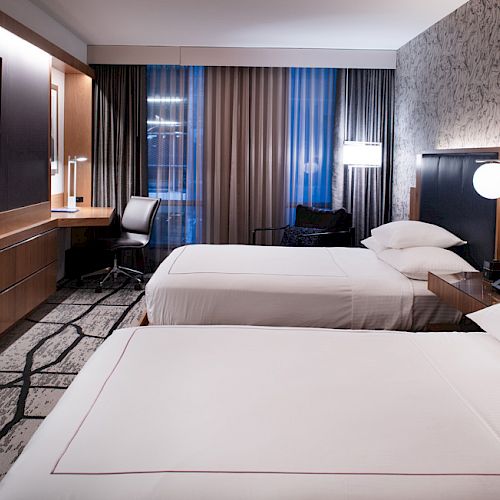 A modern hotel room with two neatly made beds, a wall-mounted TV, a wooden desk, and a large window with curtains.