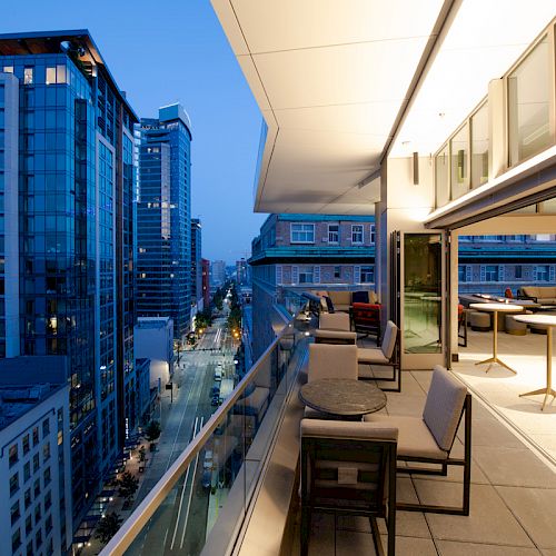 A modern cityscape at twilight with high-rise buildings, viewed from a balcony featuring seating areas and tables, leading into an indoor space.