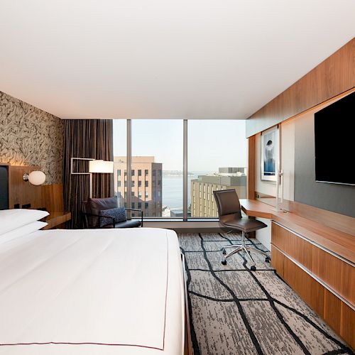 A modern hotel room features a large bed, desk, chair, and flat-screen TV with a view of buildings and water through floor-to-ceiling windows.