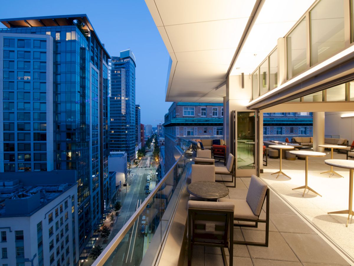 A modern balcony with outdoor seating overlooks a city street lined with tall buildings at twilight. The indoor area has tables and chairs.