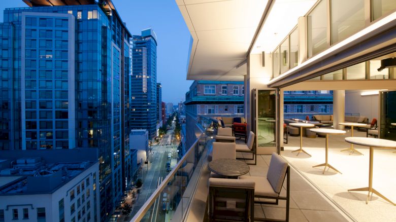 A modern balcony with outdoor seating overlooks a city street lined with tall buildings at twilight. The indoor area has tables and chairs.