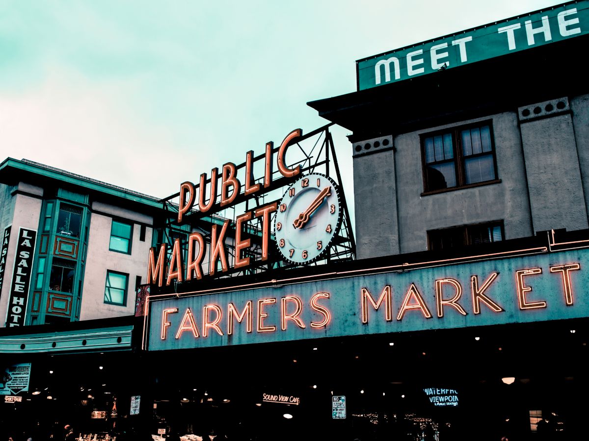 The image shows a public market with a clock sign reading "Public Market" and "Farmers Market," surrounded by buildings with various signs.
