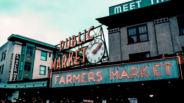 The image shows a public market with a clock sign reading "Public Market" and "Farmers Market," surrounded by buildings with various signs.