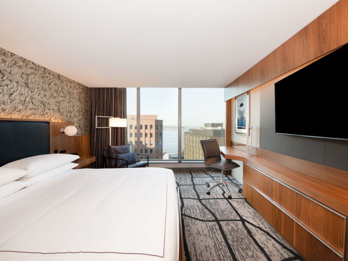 A modern hotel room with a large bed, desk, chair, wall-mounted TV, and floor-to-ceiling window offering a city and partial water view.