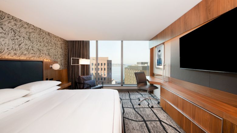 A modern hotel room with a large bed, desk, chair, wall-mounted TV, and floor-to-ceiling window offering a city and partial water view.