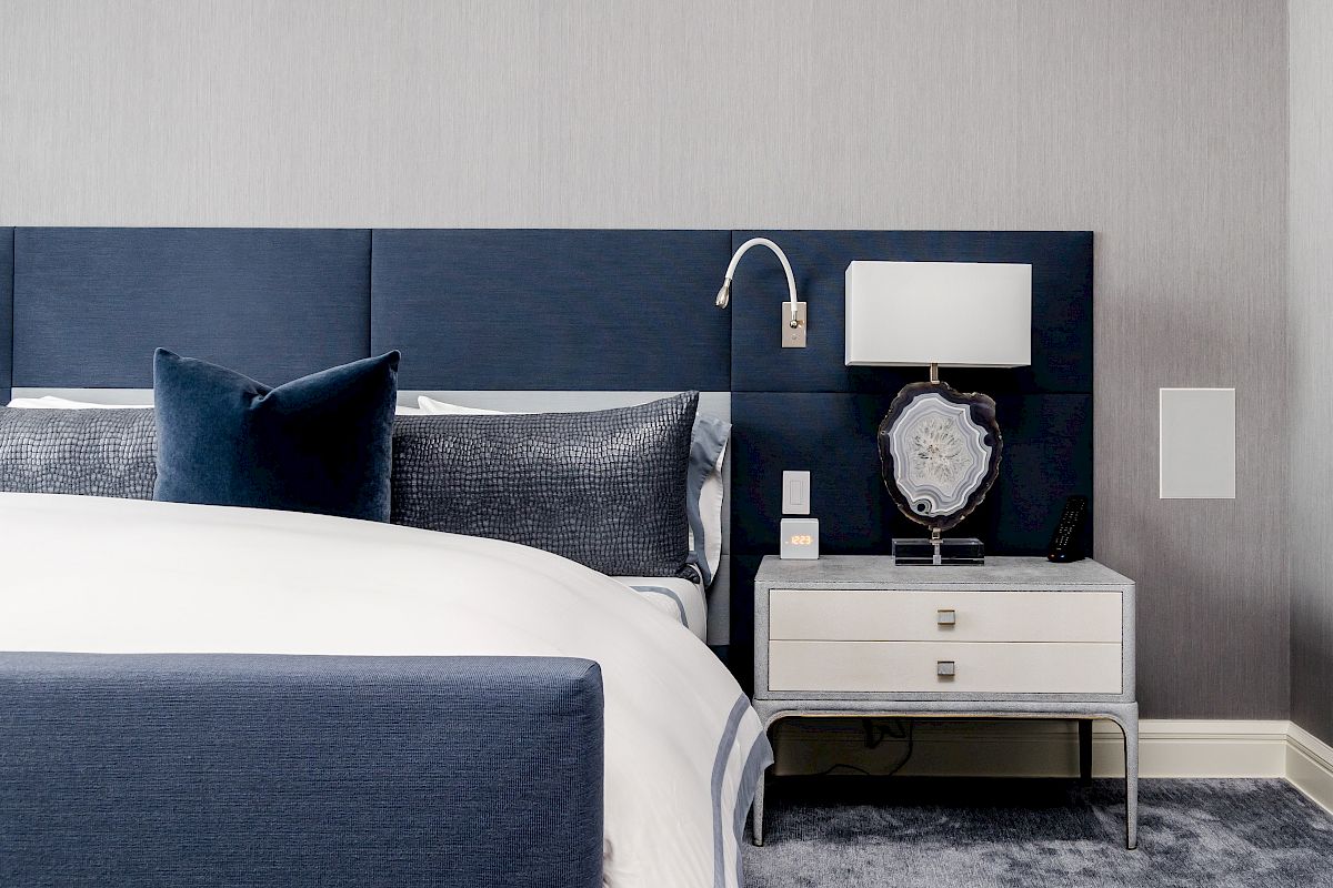 This image shows a modern bedroom with a blue headboard, blue and gray bedding, a white nightstand with a lamp, and a neutral wall background.