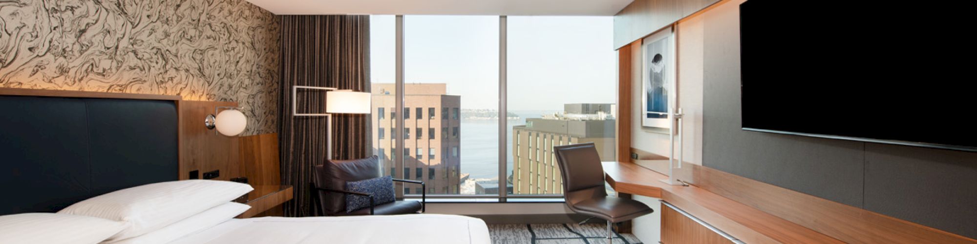 A modern hotel room with a large bed, a desk, a chair, a flat-screen TV, and a window with a view of city buildings and water.
