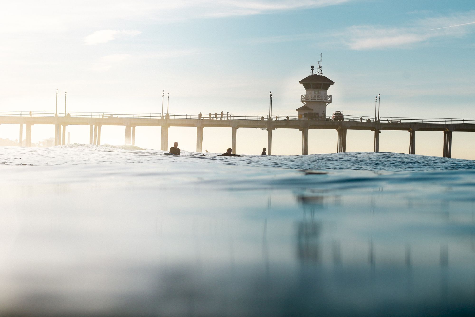 The image shows a scenic view of a pier extending over the water, with several people on it and a few surfers in the foreground.